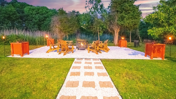 An outdoor seating area with wooden furniture and a fire pit, surrounded by trees and a gravel path.