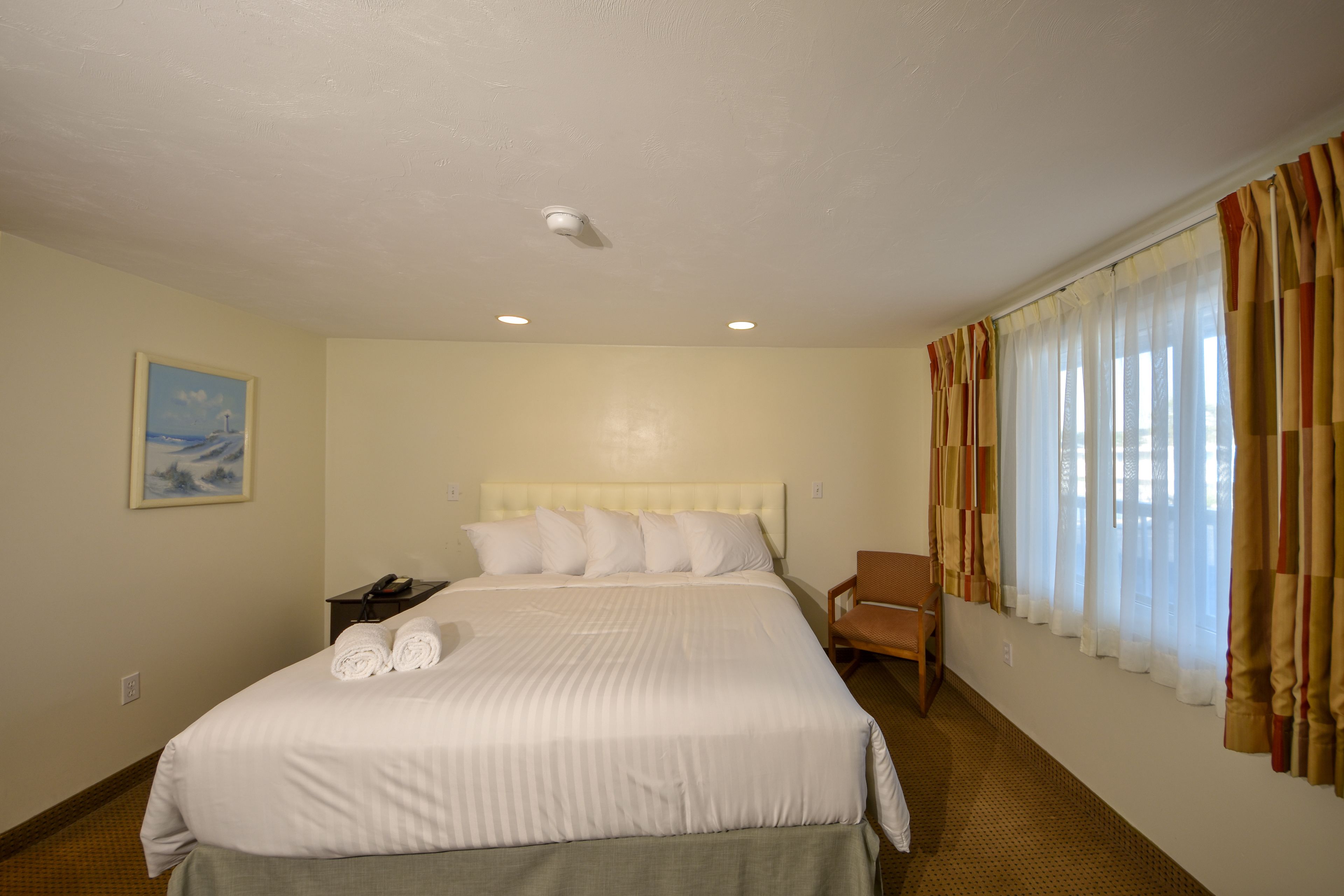 Una habitación de hotel con una cama grande, cabecera, mesita de noche, una silla y un ventanal con cortinas.