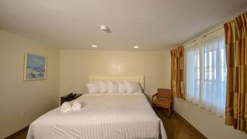 Una habitación de hotel con una cama grande, cabecera, mesita de noche, una silla y un ventanal con cortinas.