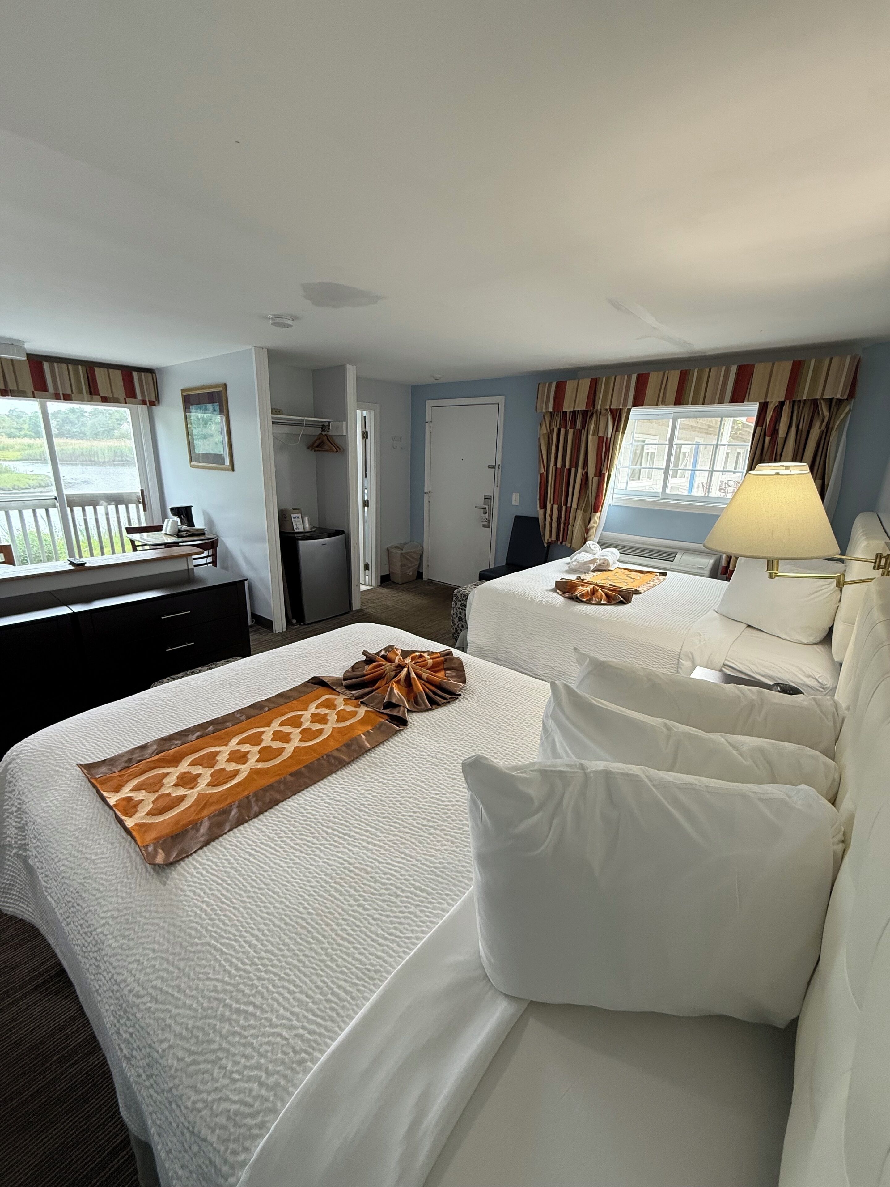 Habitación de hotel con dos camas, un escritorio, una silla, una lámpara y vista a un cuerpo de agua a través de una ventana.
