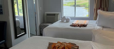 A hotel room with two beds, a large window with a view of a lake, a mirror, and a desk with a lamp.