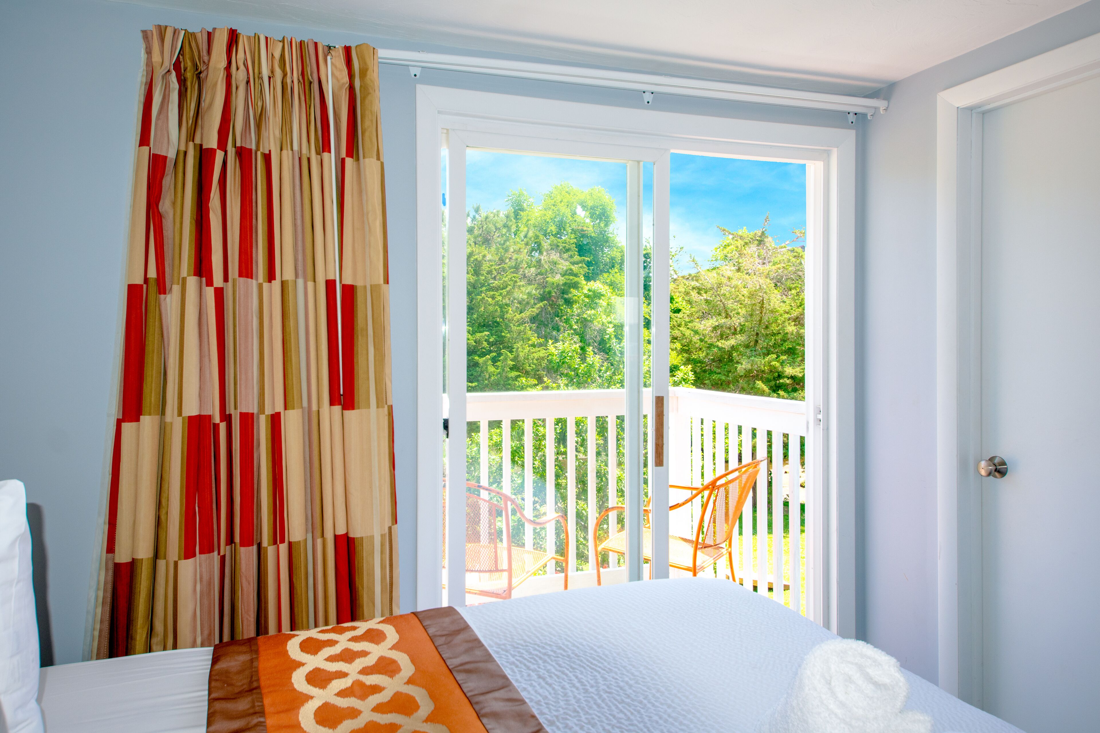 Quarto com uma cama, vista para uma varanda com cadeiras e uma janela com cortinas.