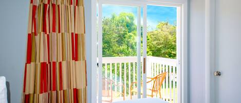 A bedroom with a bed, a view of a balcony with chairs, and a window with curtains.