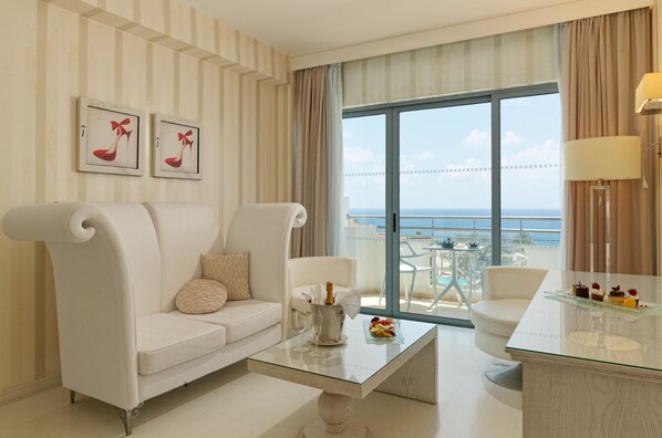 Una habitación de hotel moderna con un sofá blanco, una mesa de centro de vidrio con una botella y vasos, un balcón con vistas al mar y un comedor con una mesa de vidrio y postre.
