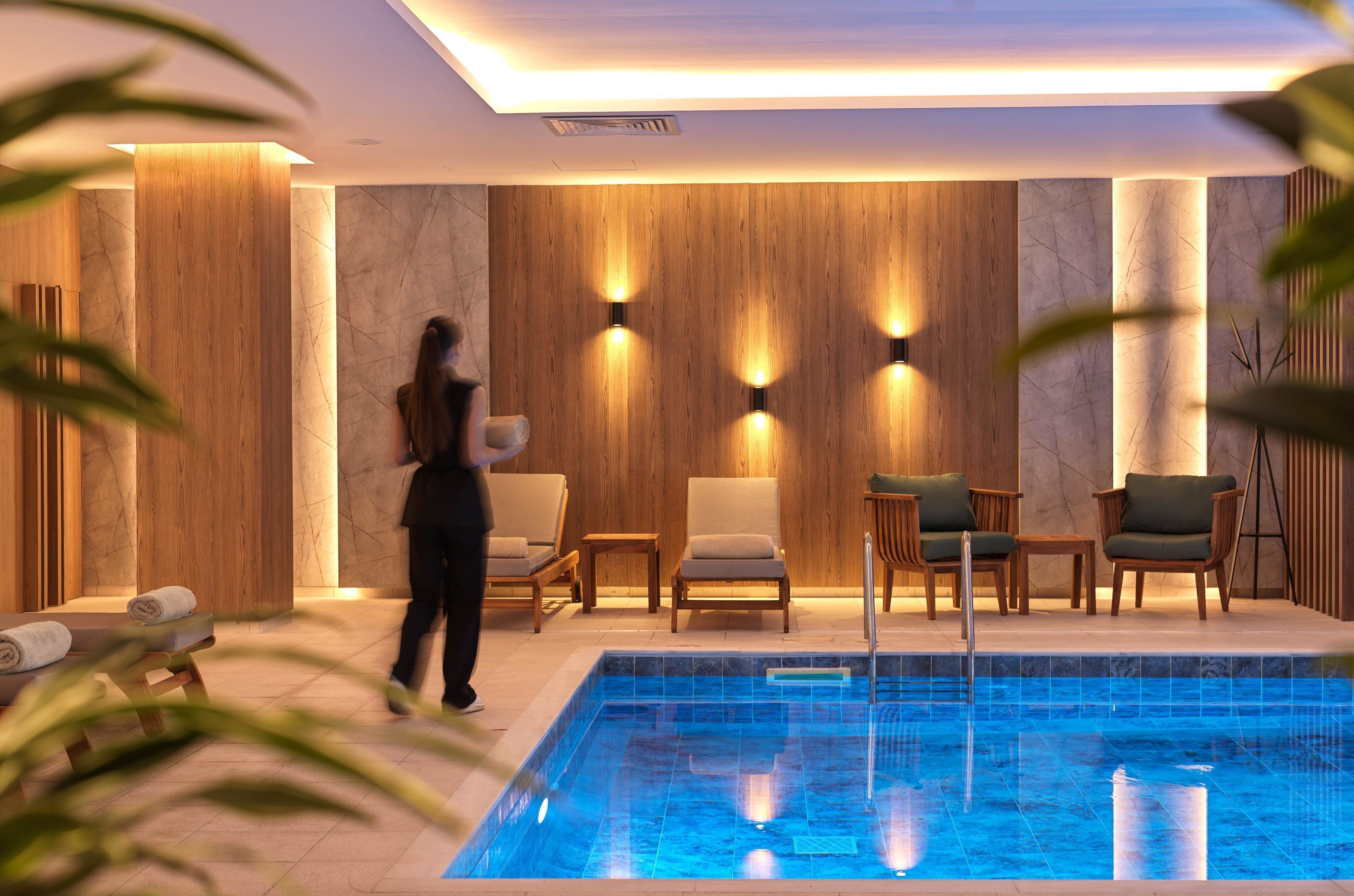 Un área de piscina interior moderna con paneles de madera, sillones de descanso y una piscina azul cristalina.