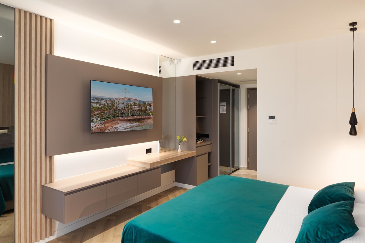 Un dormitorio moderno con una cama, una mesita de noche, un espejo y un televisor de pared.