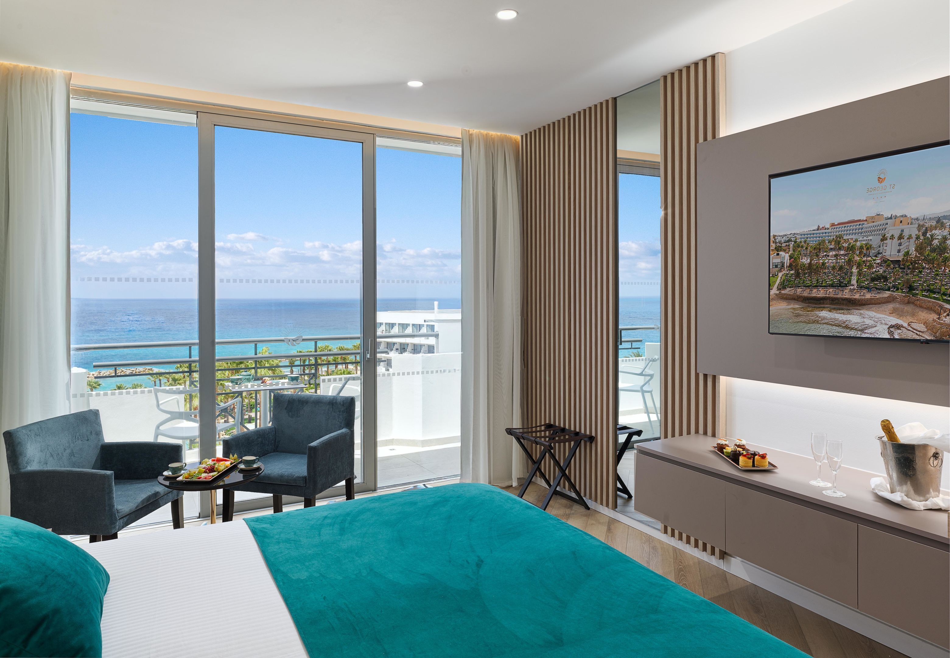 A hotel room with a large balcony, a bed with teal bedding, a dining table with a tray of food, and a minibar with champagne glasses.