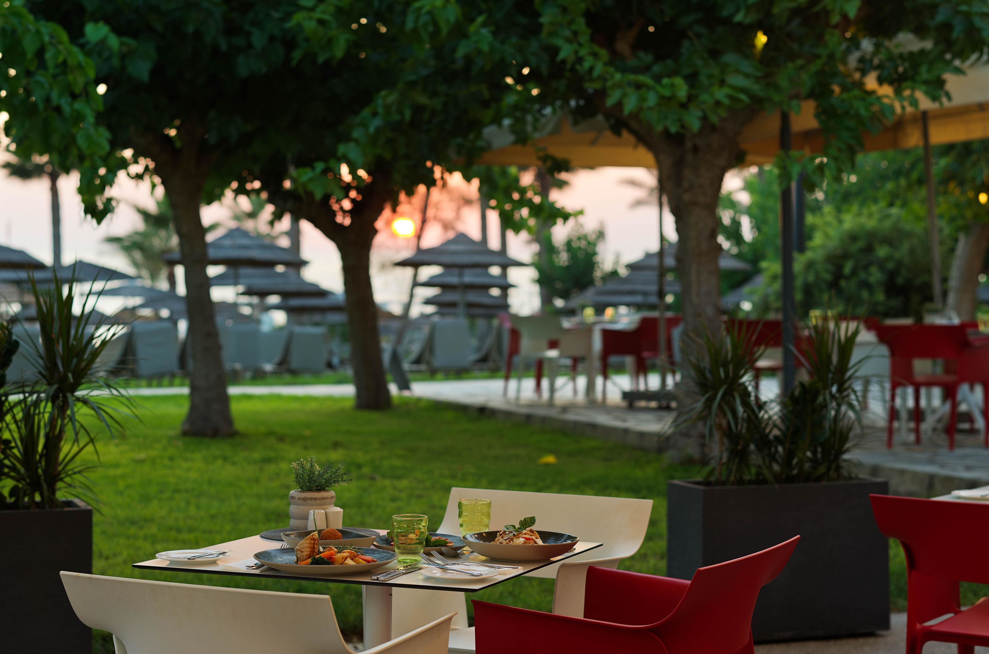 An outdoor dining area with tables and chairs, surrounded by greenery and umbrellas.
