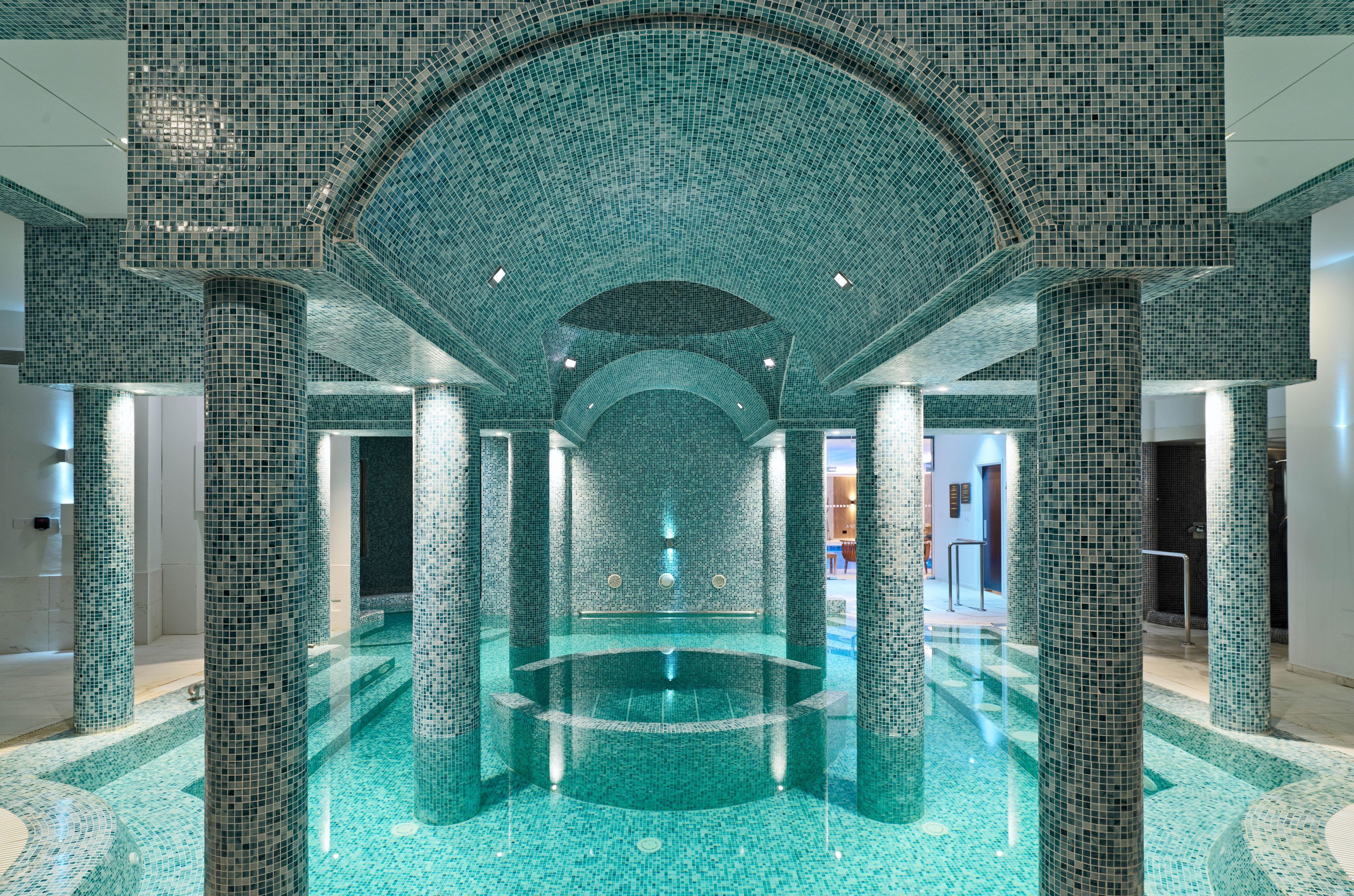 En stor indendørs swimmingpool med vægge og søjler af mosaikfliser, omgivet af et flisegulv.