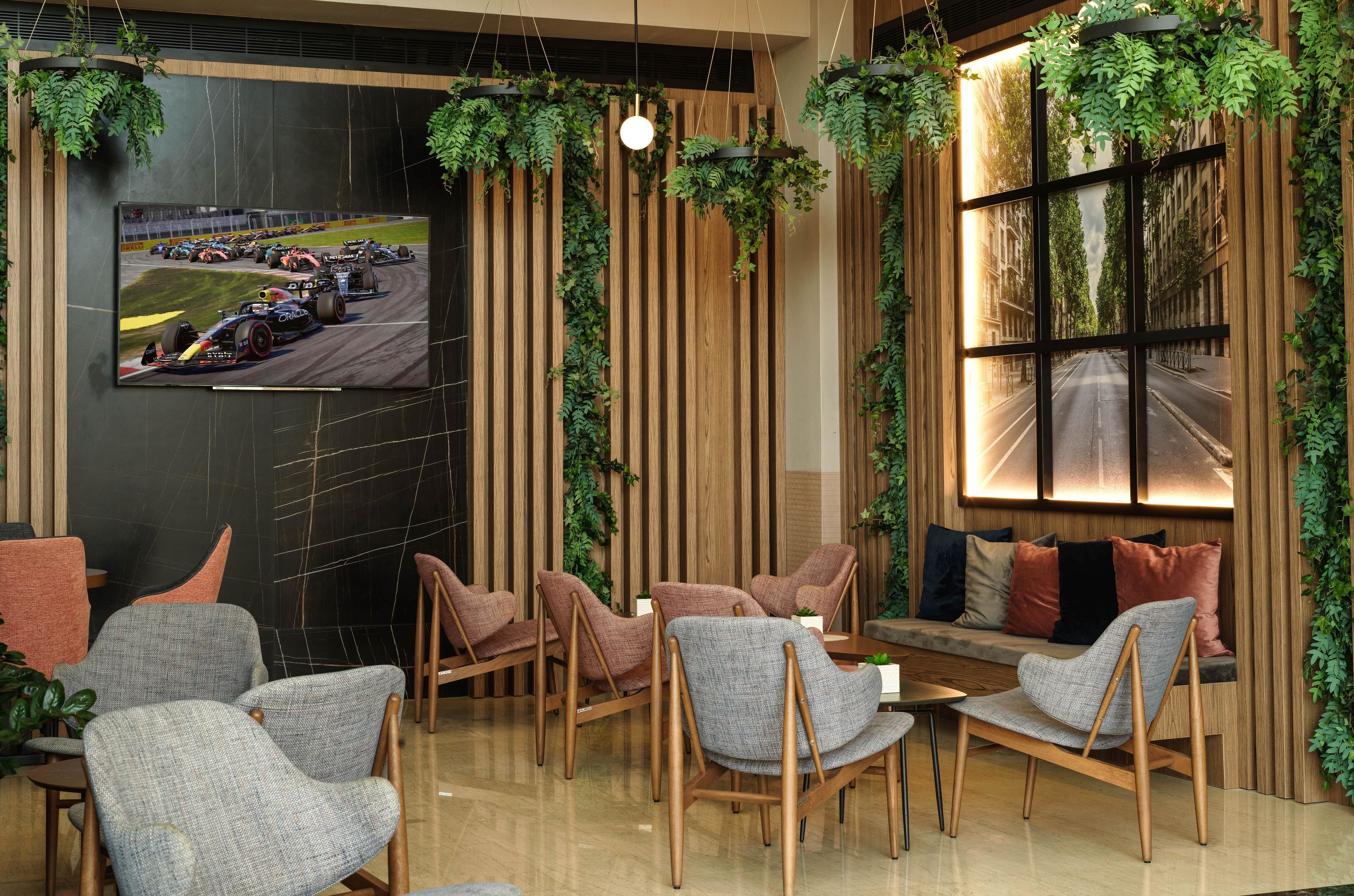 A modern lounge area with seating, a television displaying a racing event, and vertical wooden panels with greenery.