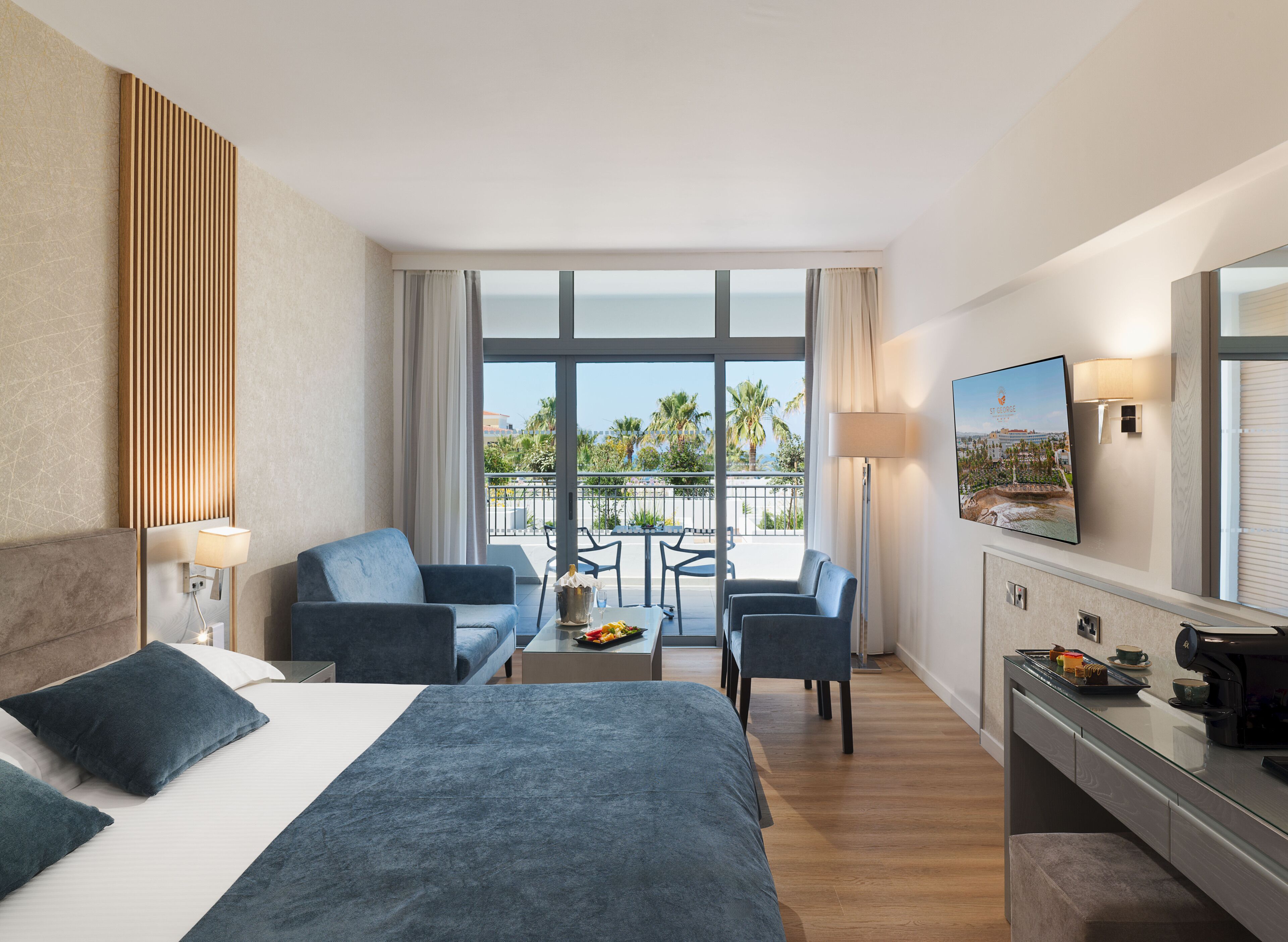 A hotel room with a bed, a sofa, a desk with a lamp, a TV, and a balcony with a view of palm trees.