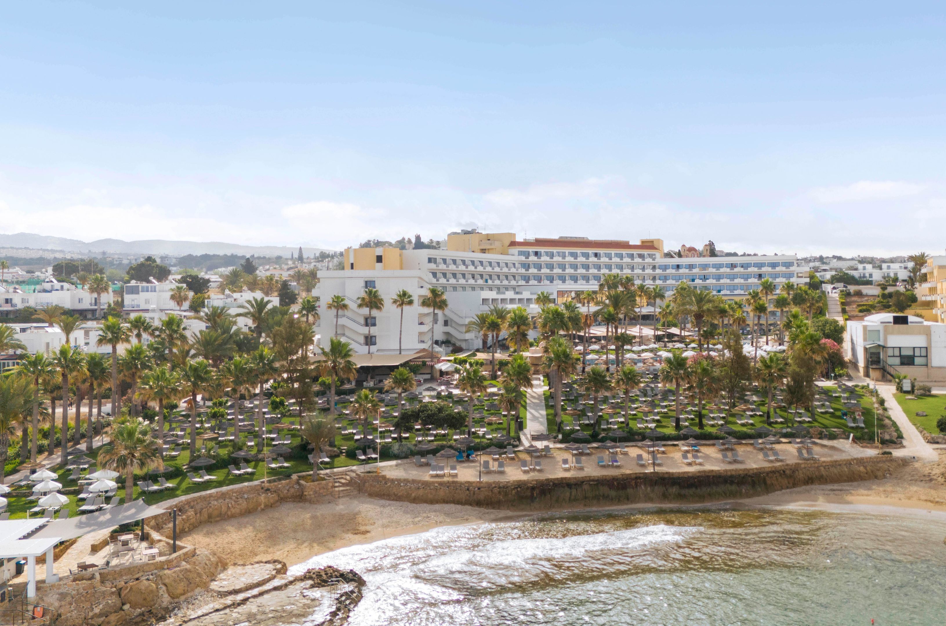 Um hotel à beira-mar com vários andares, palmeiras e uma área de praia com espreguiçadeiras.