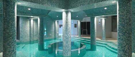 Una amplia piscina interior con suelo y paredes de azulejos, rodeada de pilares y columnas.