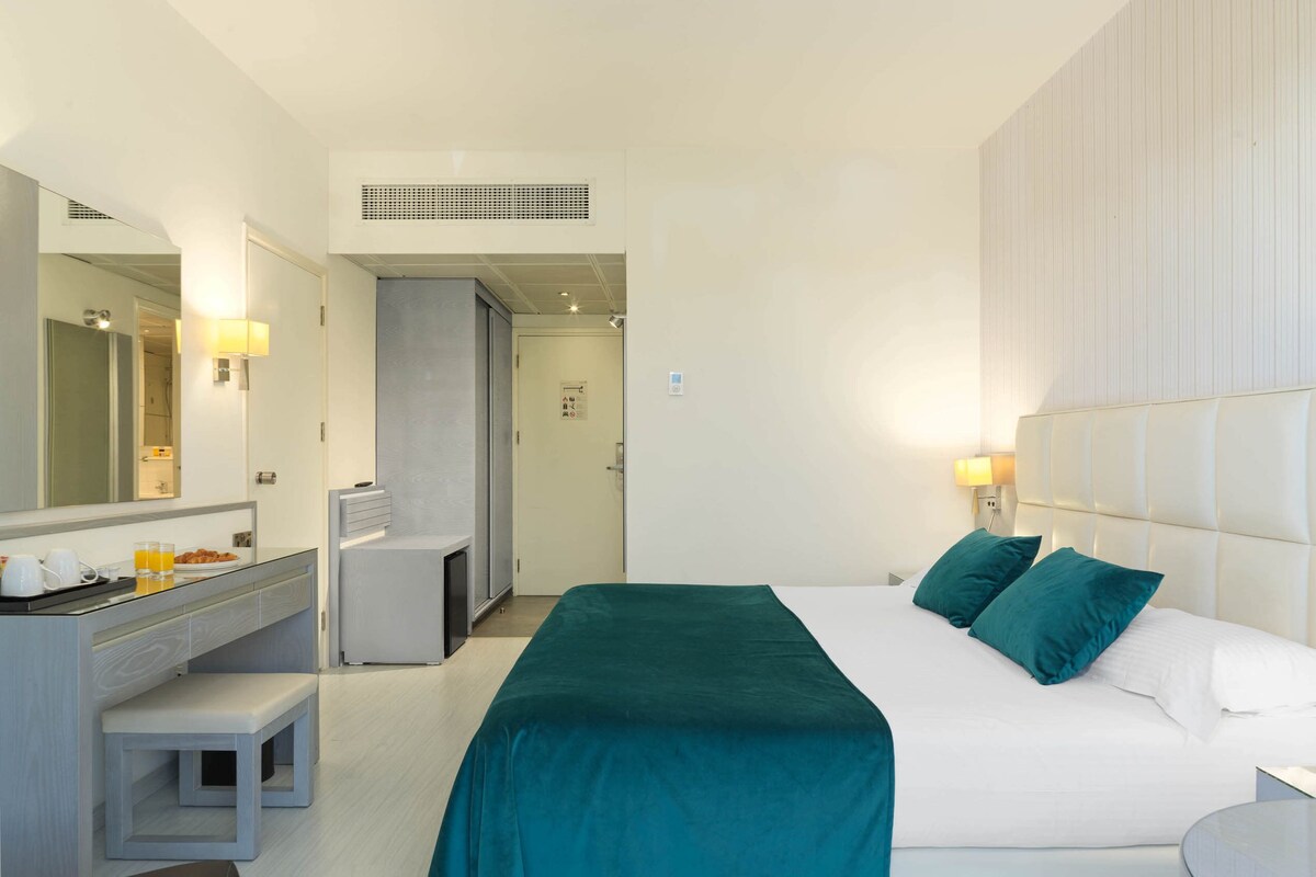 Una habitación de hotel con cama, escritorio con silla, espejo y una pequeña kitchenette.