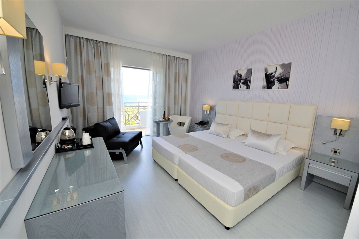 Habitación de hotel moderna con una cama grande, un escritorio, una silla, un televisor y un balcón con vistas.
