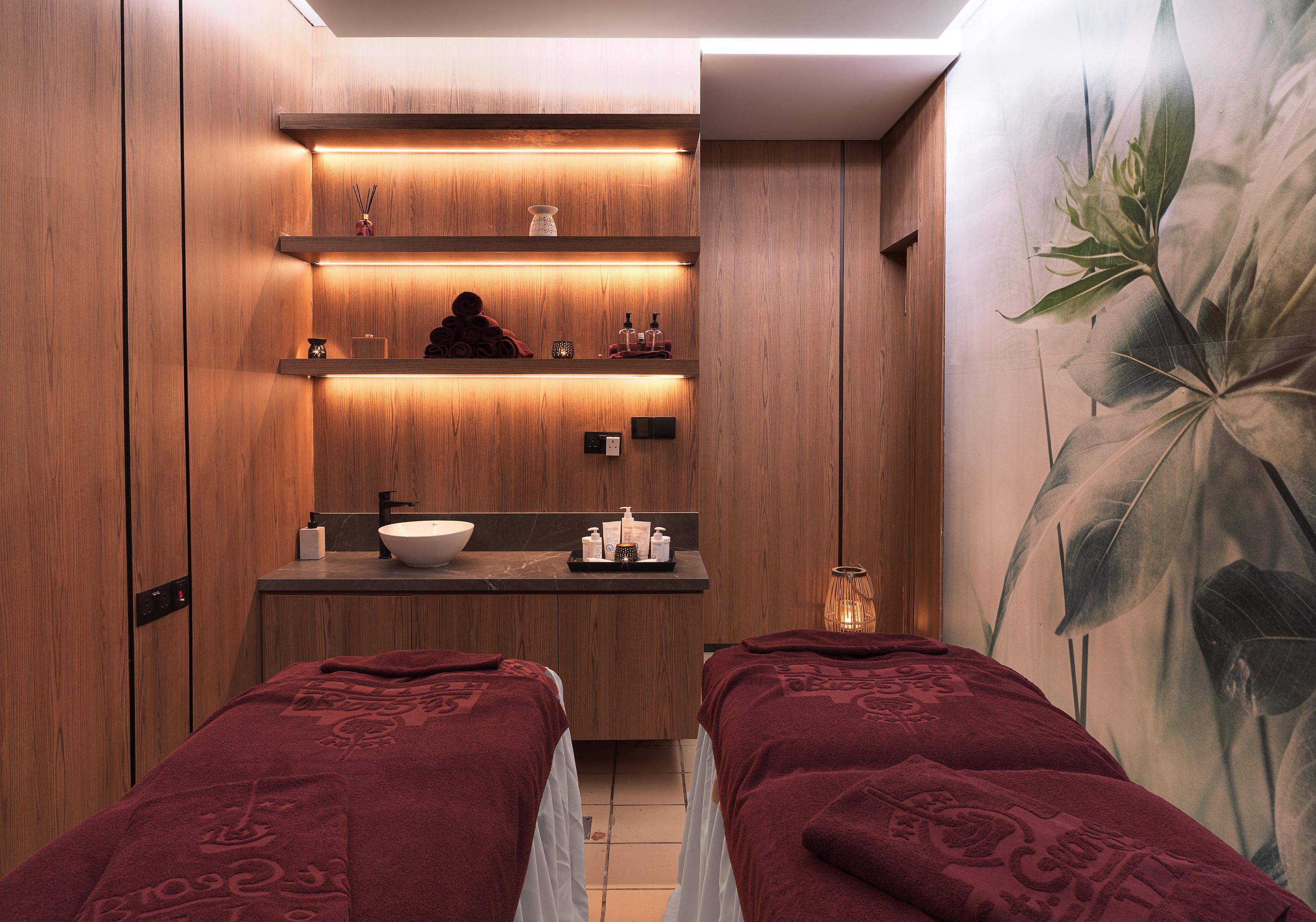 A spa room with wooden paneling, a large mirror, a sink, and two beds with red towels.