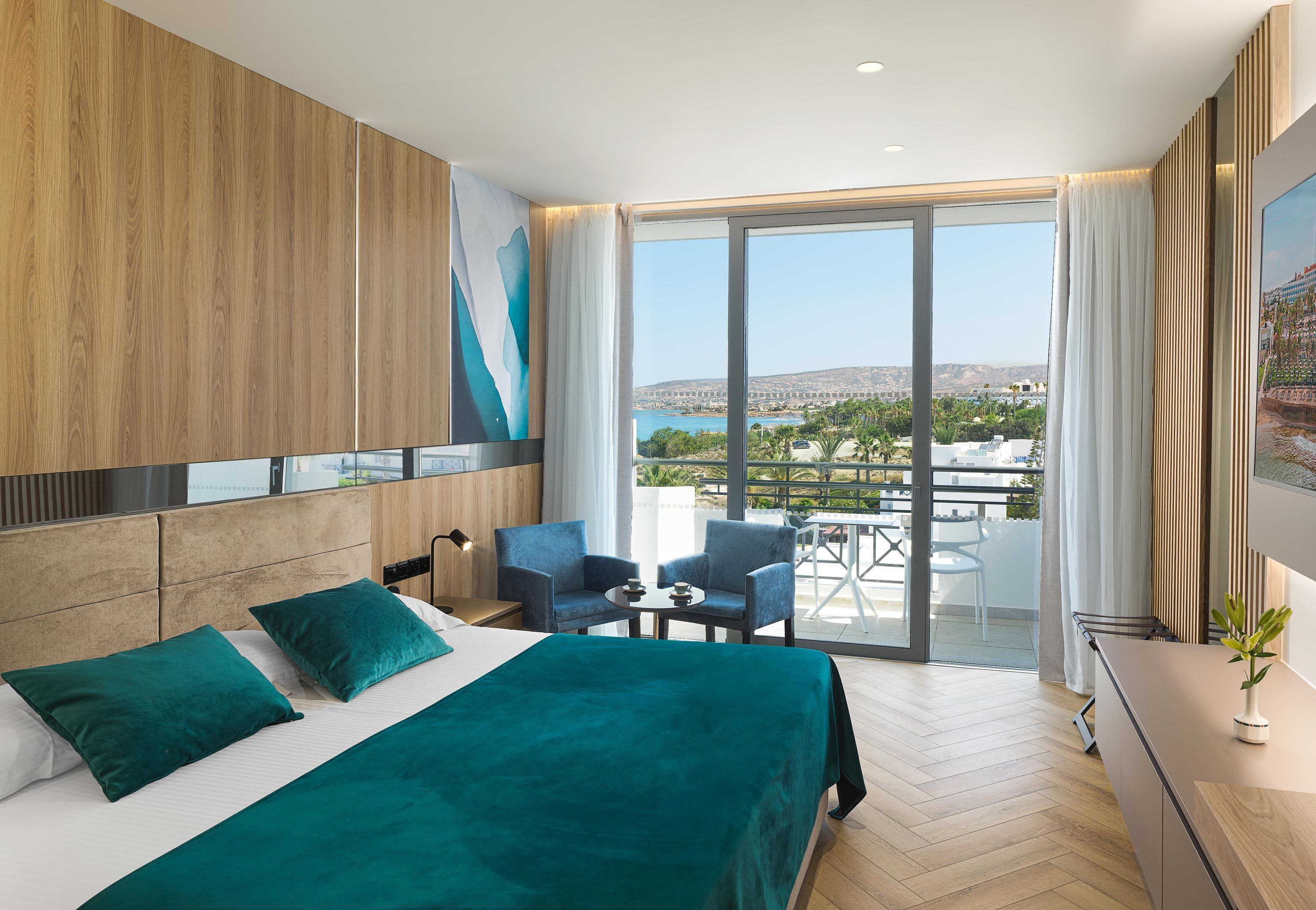 Une chambre d’hôtel moderne avec un grand lit, deux fauteuils, une petite table et un balcon offrant une vue sur la ville.