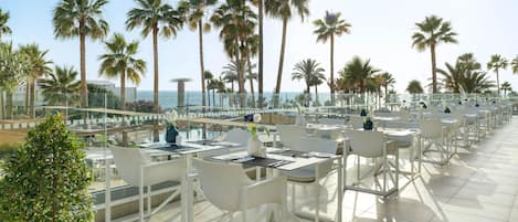Ein Dachterrassenrestaurant mit weißen Tischen und Stühlen, mit Blick auf einen Strand mit Palmen und das Meer.
