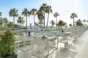 Un área de comedor en la azotea con mesas y sillas blancas, con vistas a una playa con palmeras y el mar.