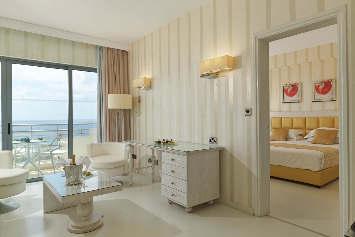 Habitación de hotel con un ventanal grande, un escritorio con superficie de vidrio, una cama con cabecero y un minibar.