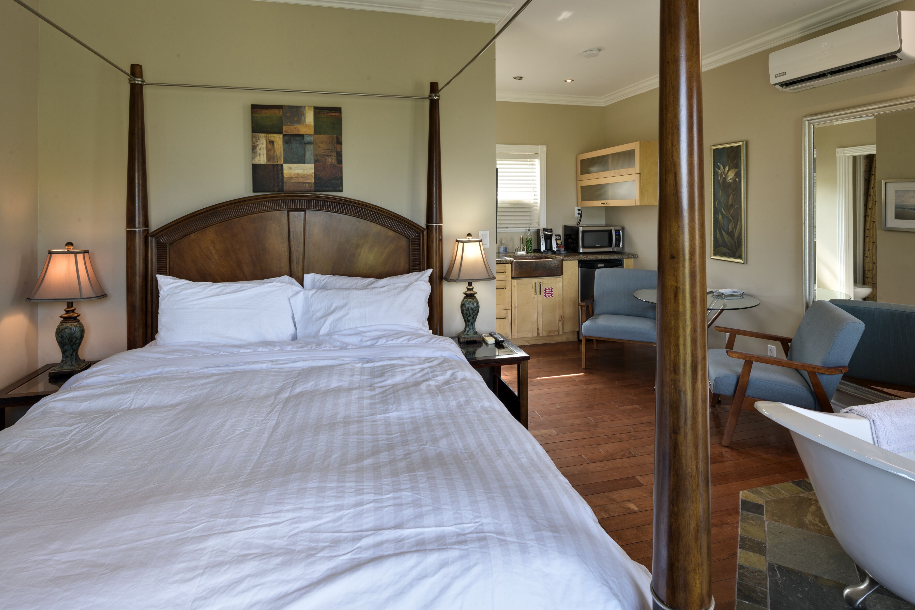 A four-poster bed with white linens, a wooden headboard, a bedside table with a lamp, a small desk with a lamp, a blue armchair, a white bathtub, and a kitchenette area with a microwave and cabinets.