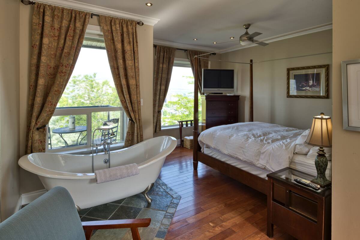 A bedroom with a large bed, a bathtub, a television, and a balcony with a table and chairs.