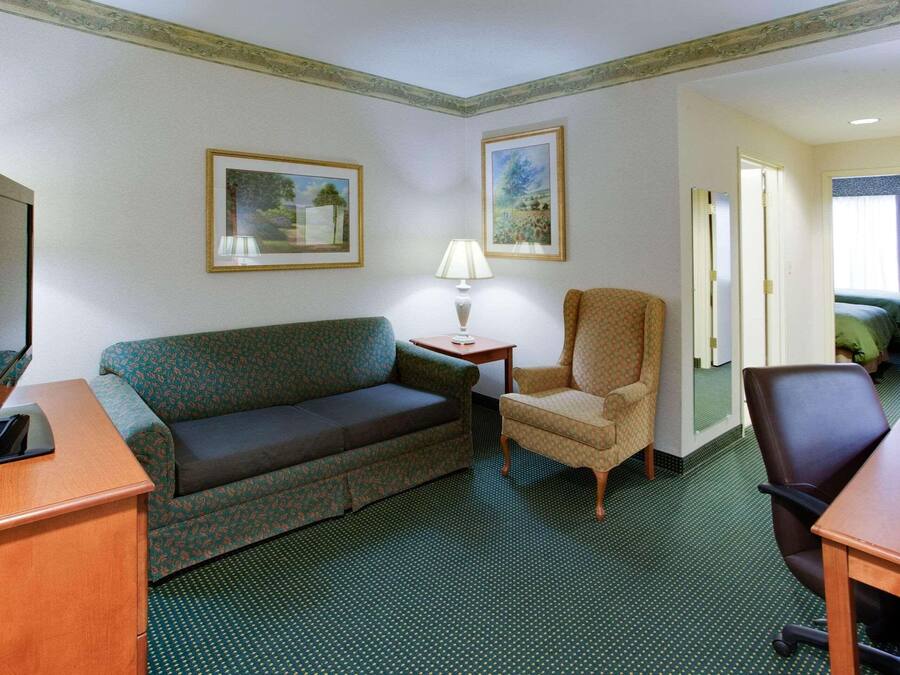Une chambre d’hôtel avec un canapé à motifs verts, un petit fauteuil, un bureau en bois avec une télévision et deux tableaux encadrés accrochés au mur.