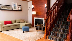 A living room with a sectional sofa, a blue ottoman, a fireplace, and a wooden staircase with patterned carpet. - Country Inn & Suites by Radisson, Brockton (Boston), MA (Brockton)