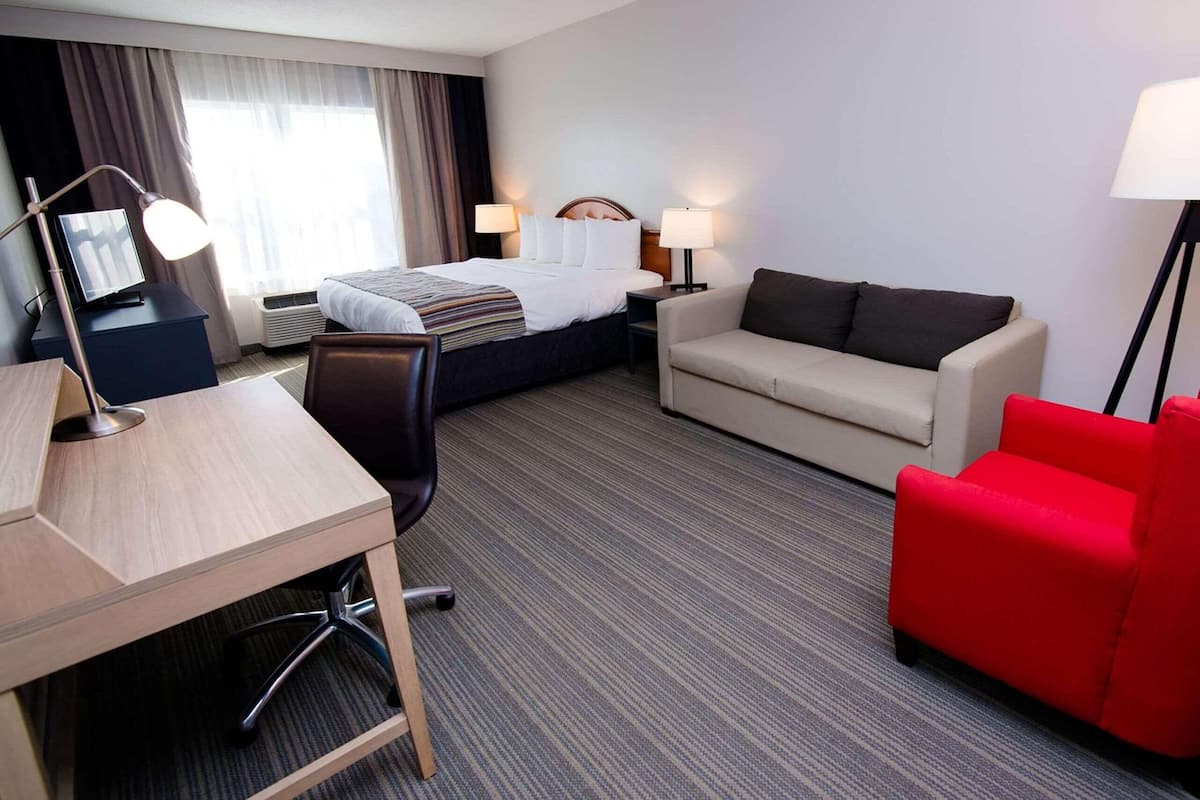 A hotel room with a bed, a desk, a red armchair, a sofa, and a standing lamp.