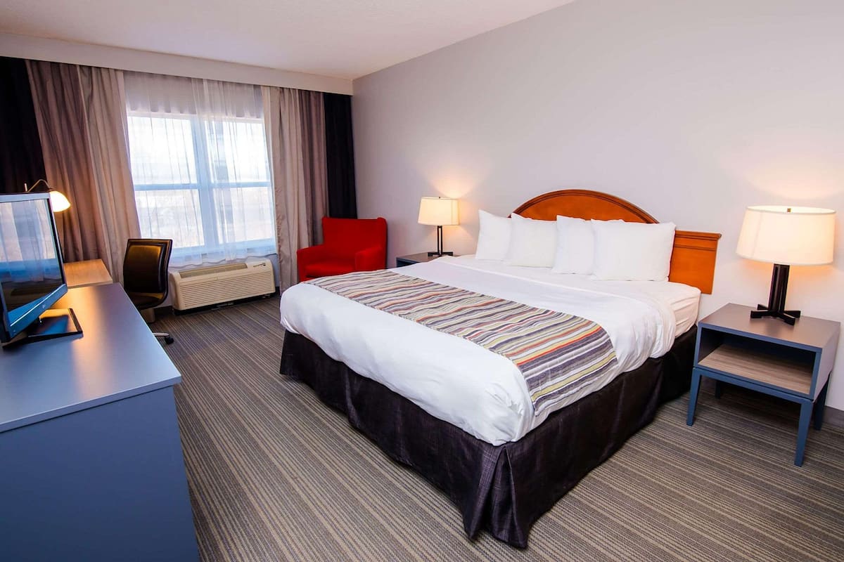 A hotel room with a large bed, two bedside tables with lamps, a desk with a computer, a red chair, and a large window with curtains.
