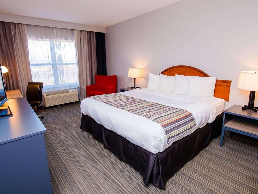 A hotel room with a large bed, two bedside tables with lamps, a desk with a computer, a red chair, and a large window with curtains.