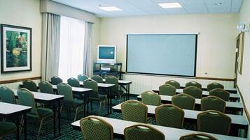A conference room with rows of tables and chairs, a projector screen, a television, and a framed picture on the wall.