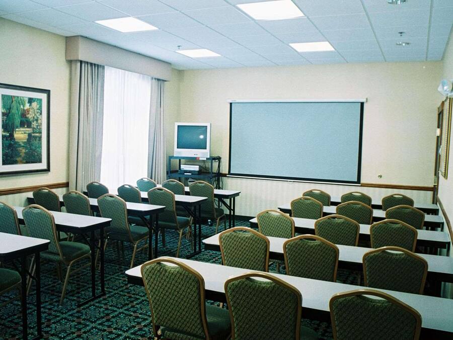 A conference room with rows of tables and chairs, a projector screen, a television, and a framed picture on the wall.