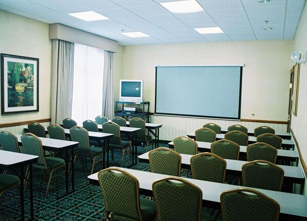 A conference room with rows of tables and chairs, a projector screen, a television, and a framed picture on the wall.