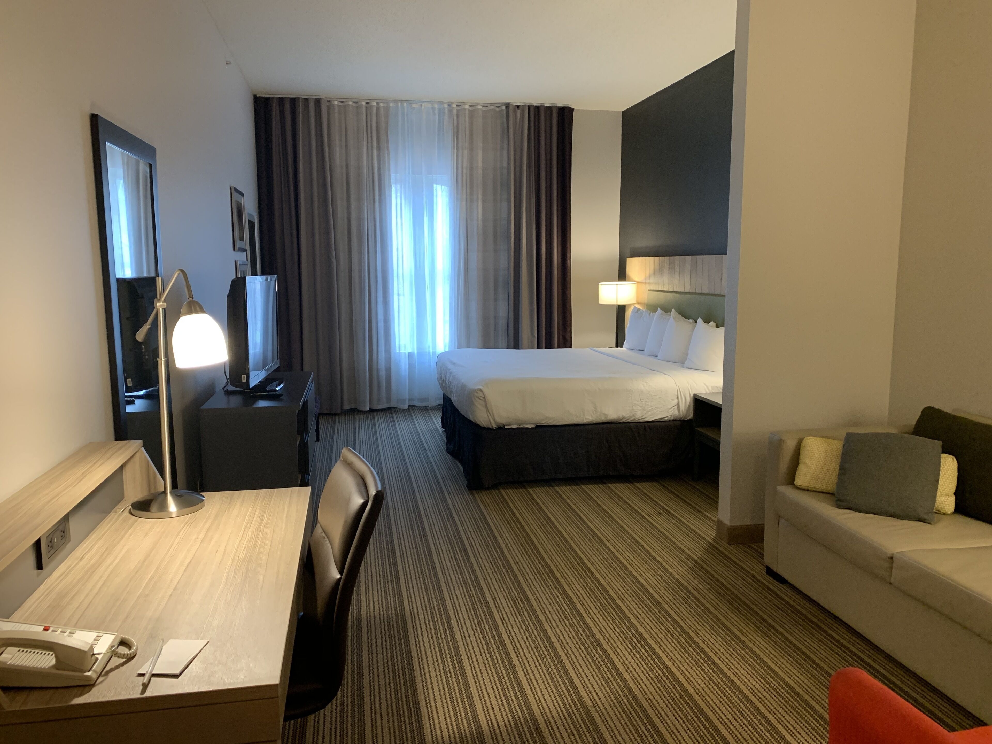 A hotel room with a bed, a desk with a telephone, a chair, a sofa, a lamp, and a large window with curtains.