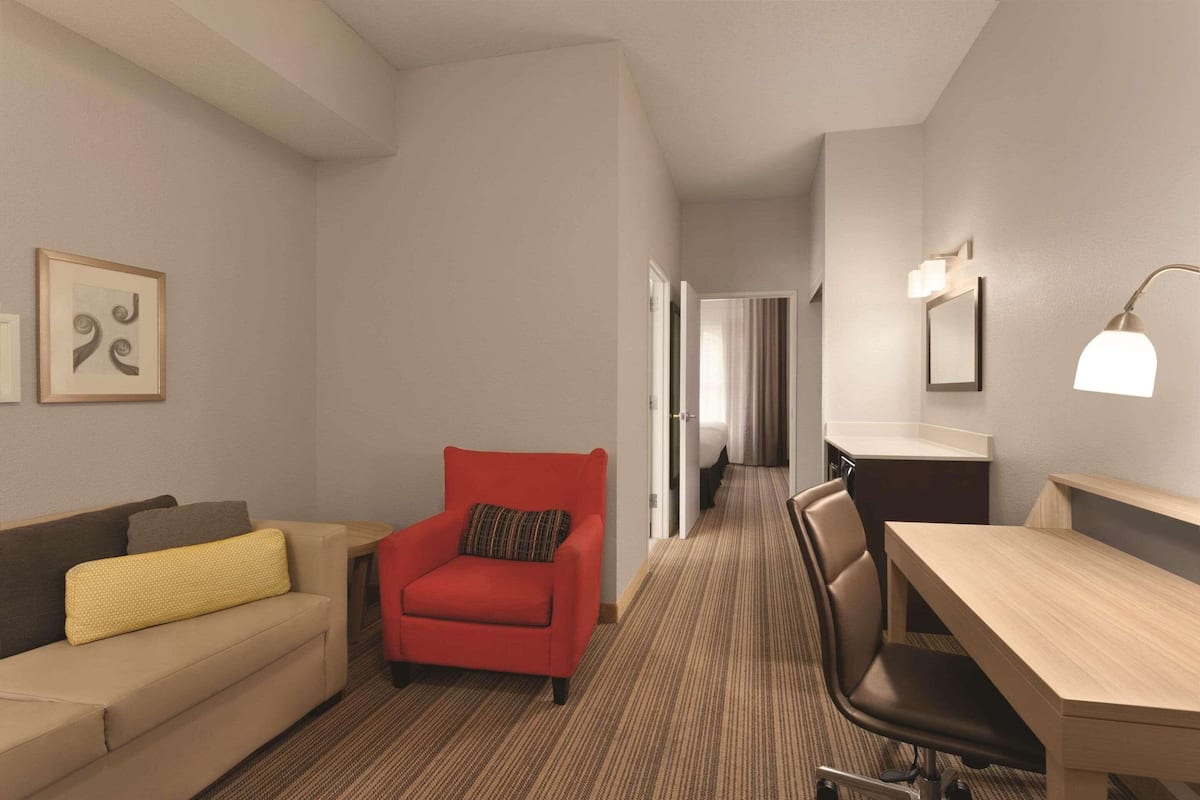 Habitación de hotel con sofá, un sillón rojo, un escritorio y una silla. Se observa un cuadro en la pared y una lámpara sobre el escritorio.