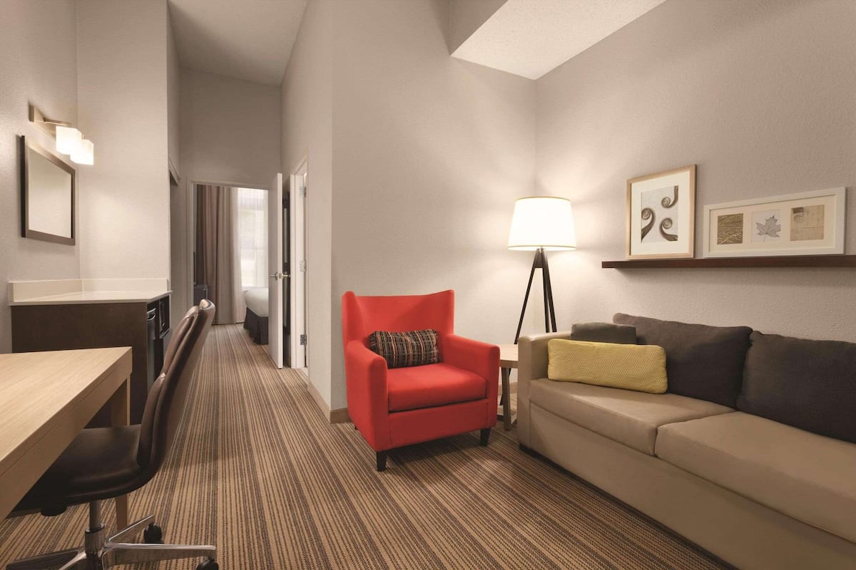 Una habitación de hotel con escritorio, un sillón rojo, un sofá, una lámpara y obras de arte enmarcadas en la pared.