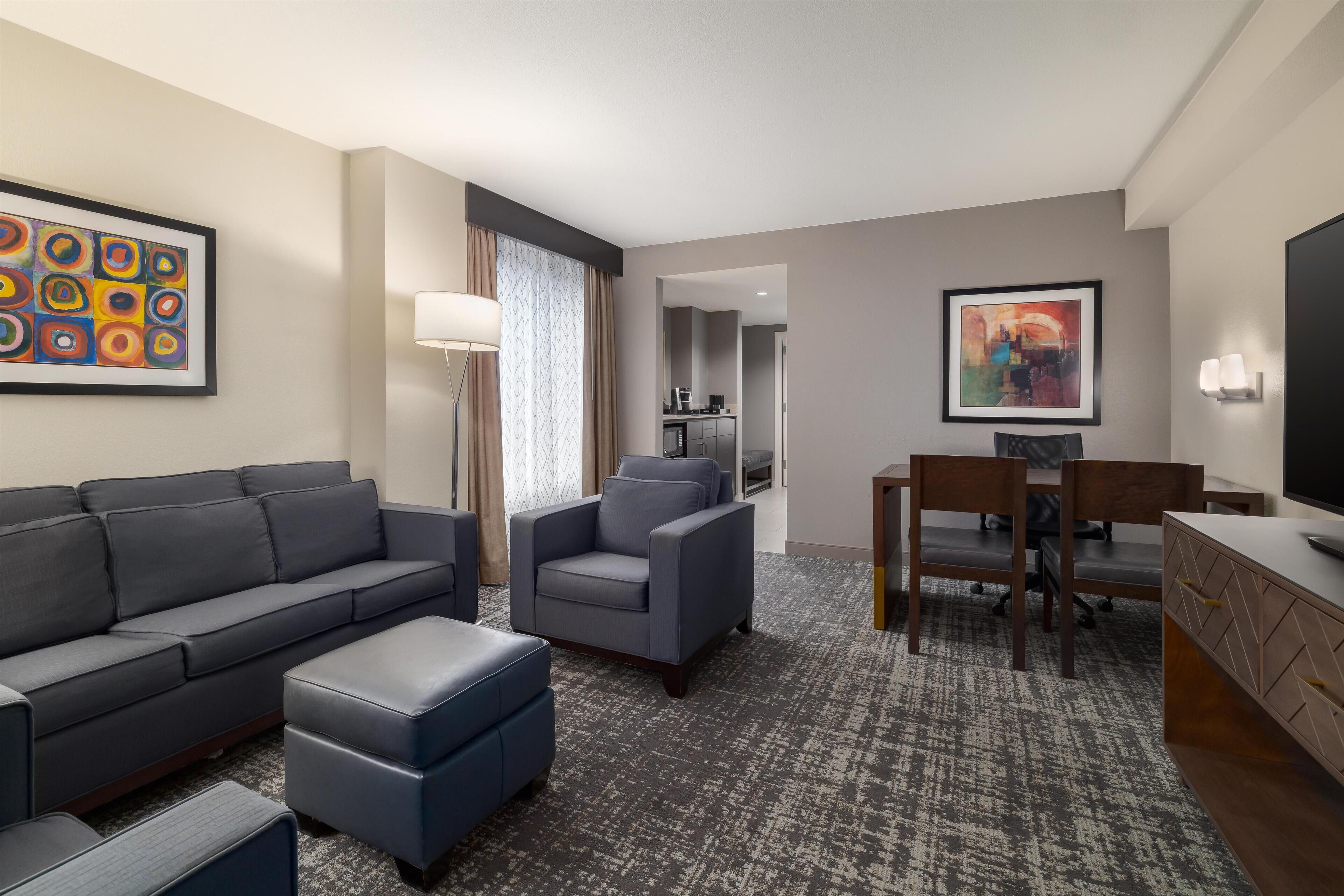 A modern hotel room with a gray sofa, a blue armchair, a small ottoman, a dining table with chairs, and a TV on a wooden cabinet.