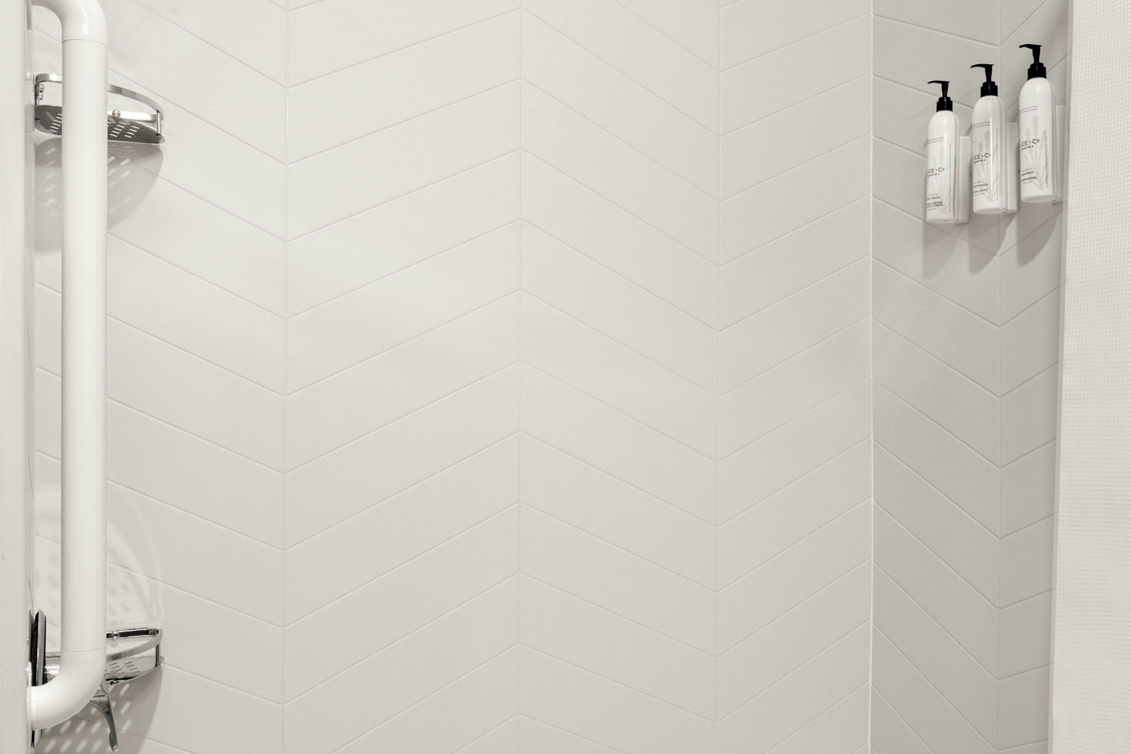 A bathroom with a white herringbone tile pattern, a shower with a chrome faucet, and three toiletry bottles on the wall.