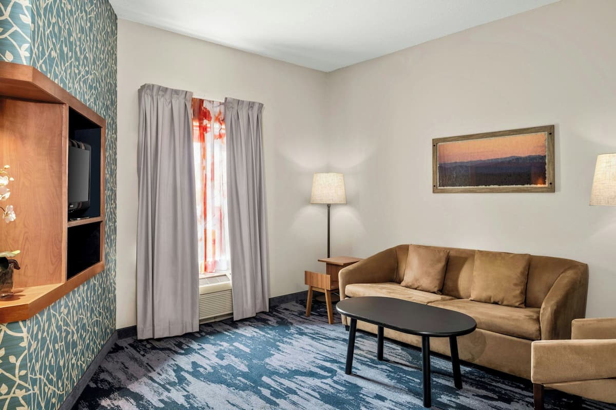 Een hotelkamer met een bank, een klein tafeltje, een lamp, een televisie op een houten plank en een raam met gordijnen.