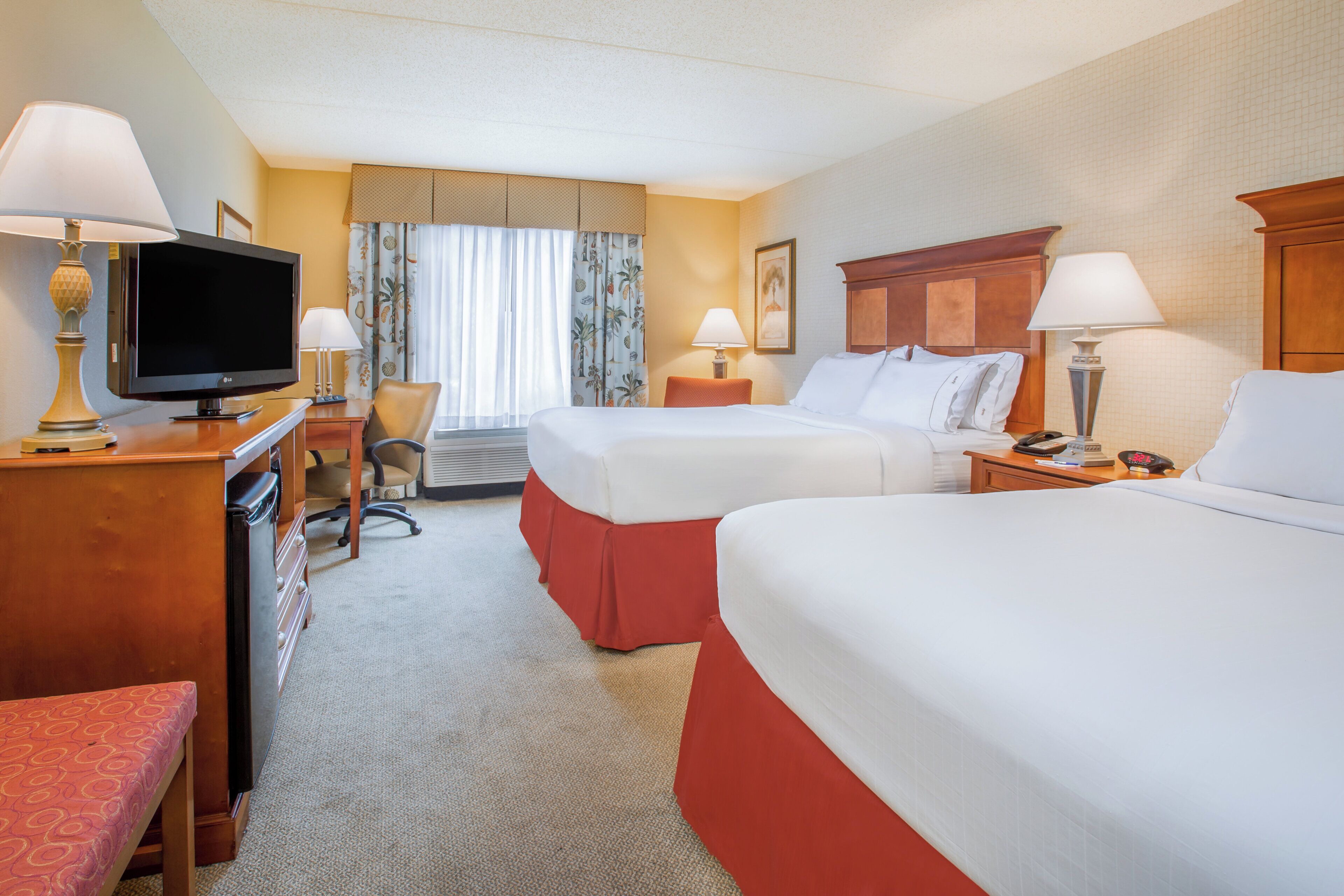 A hotel room with two beds, a desk with a TV, a chair, and a window with curtains.