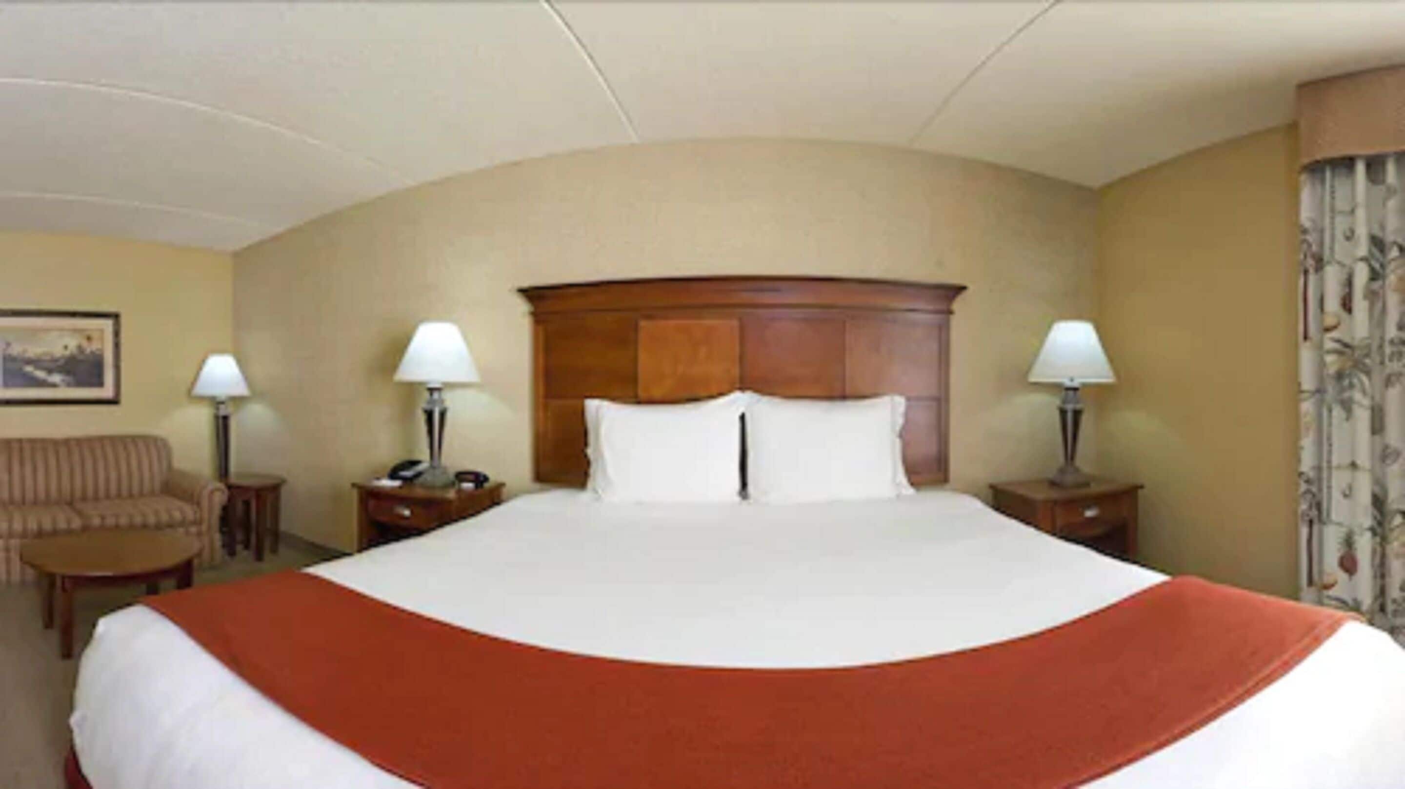 A hotel room with a large bed, two bedside tables with lamps, a wooden headboard, a sofa, and a small table.