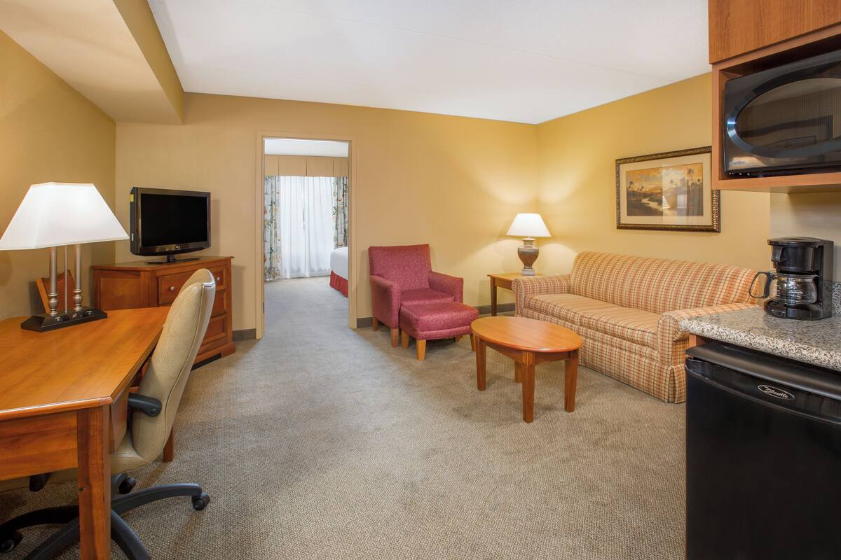 Ein Hotelzimmer mit einem Holztisch, einem Fernseher, einem roten Sessel, einem beigen Sofa, einem kleinen Tisch, einer Lampe, einer Kaffeemaschine und einer Mikrowelle.
