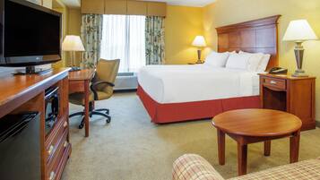 A hotel room with a bed, a desk with a television, a chair, a bedside table with a lamp, and a small round table.