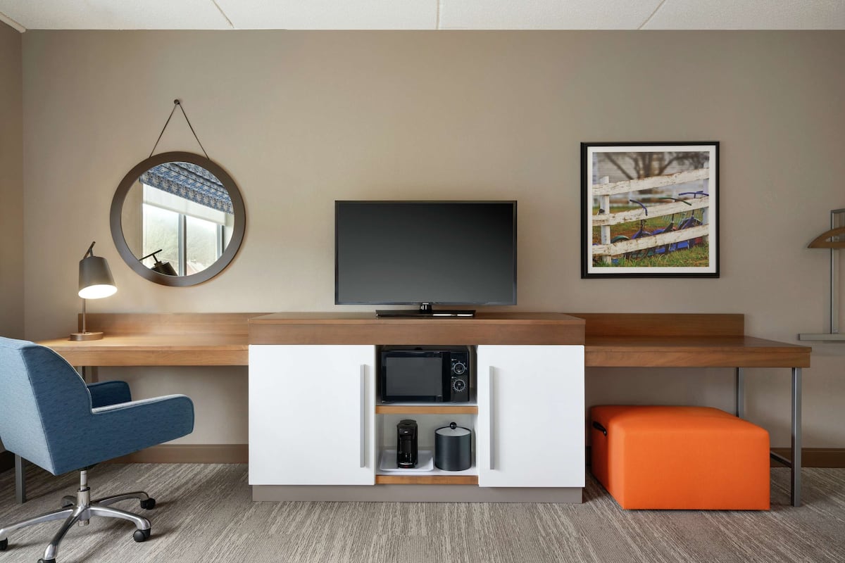 A modern hotel room with a wooden desk, a flat-screen TV, a round mirror, a framed picture, a blue office chair, a microwave, a coffee maker, and an orange ottoman.
