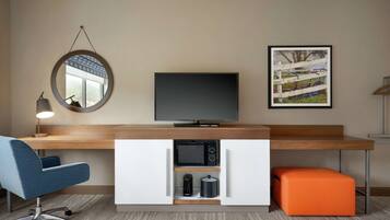 Moderni hotellihuone, jossa on puinen työpöytä, litteänäyttöinen televisio, pyöreä peili, kehystetty taulu, sininen toimistotuoli, mikroaaltouuni, kahvinkeitin ja oranssi rahi.
