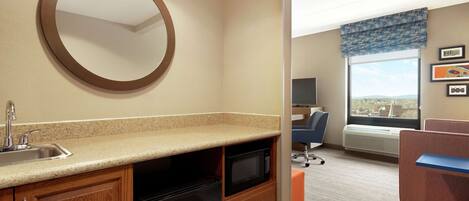 A compact hotel room with a kitchenette, a round mirror, a window with a view, and a seating area with a chair and a small table.