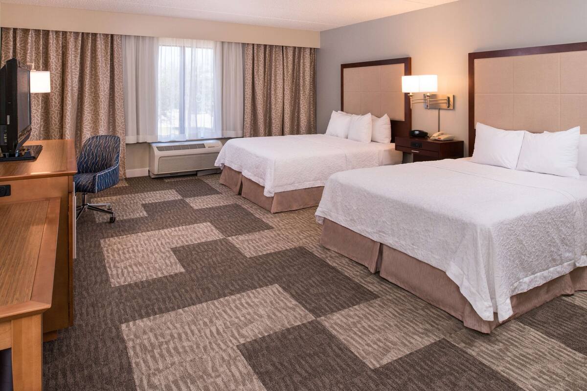 A hotel room with two beds, a desk, a chair, a TV, and a lamp.