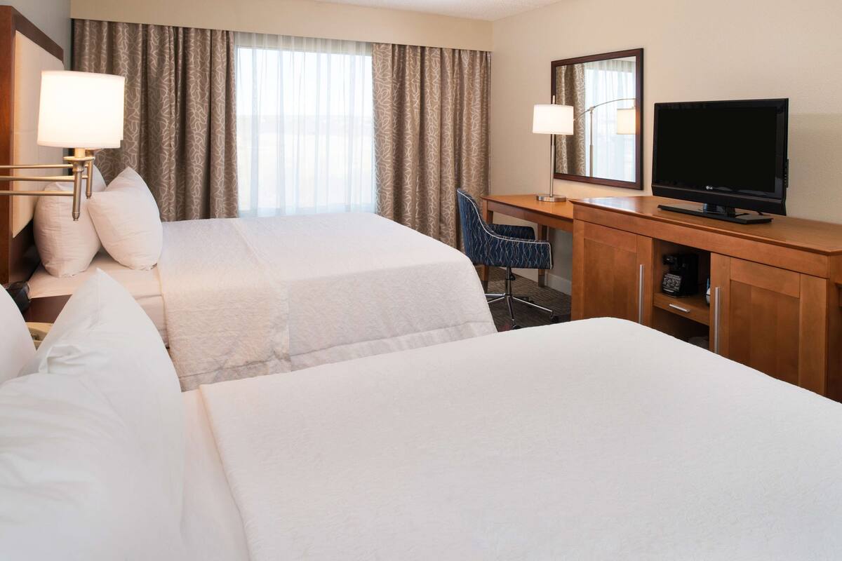 A hotel room with two beds, a desk with a TV, a chair, and a lamp.