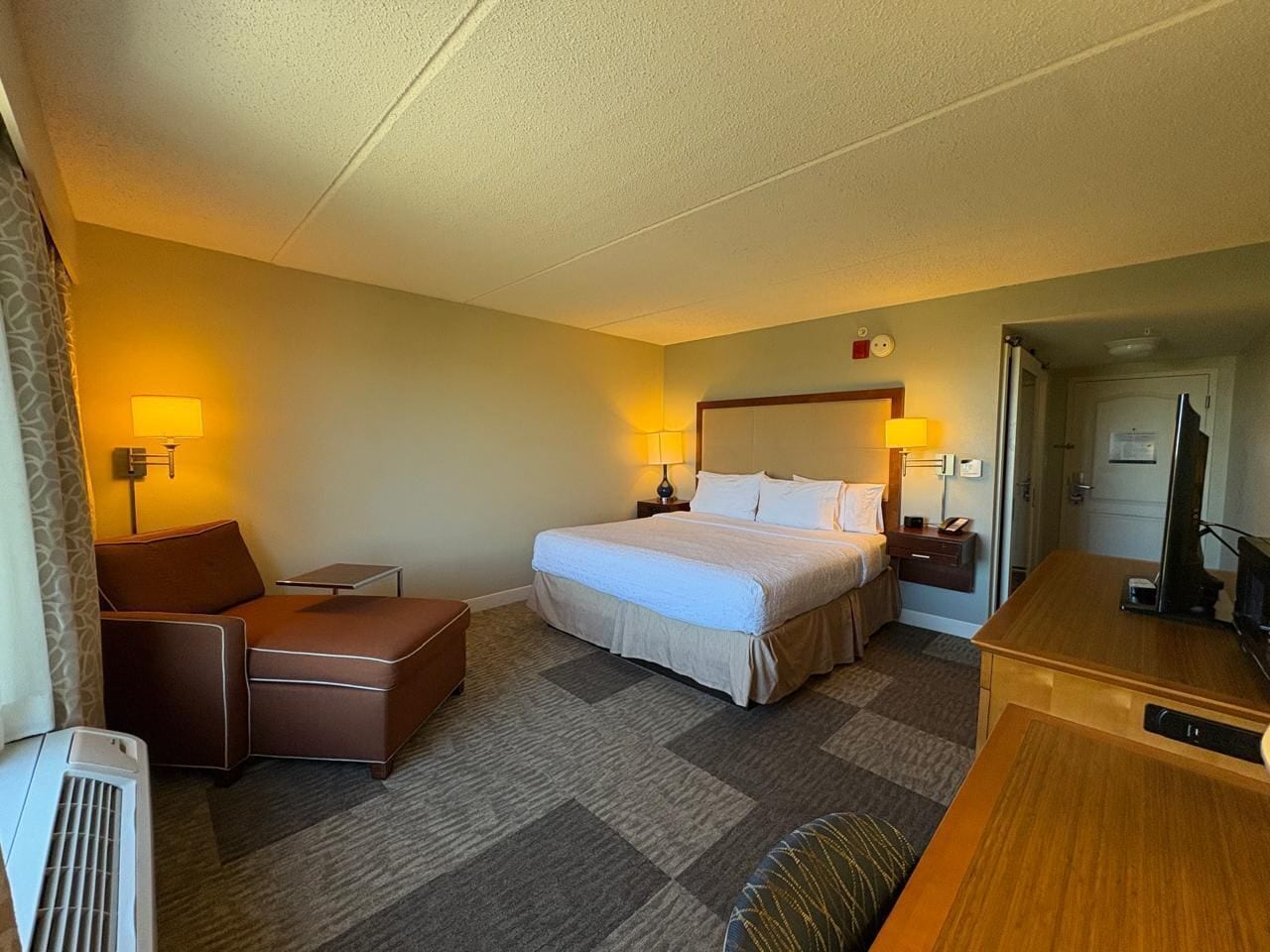 A hotel room with a bed, a desk, a chair, a small table, and a heater.